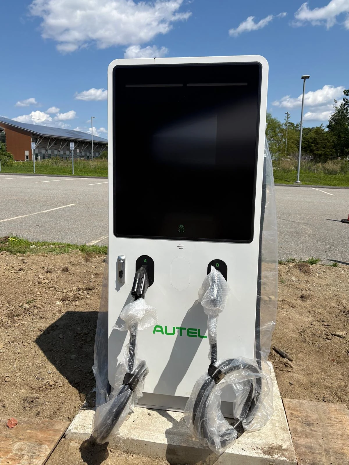 A white dual-connector DC fast charger standing on a new concrete pad, wrapped in protective plastic.