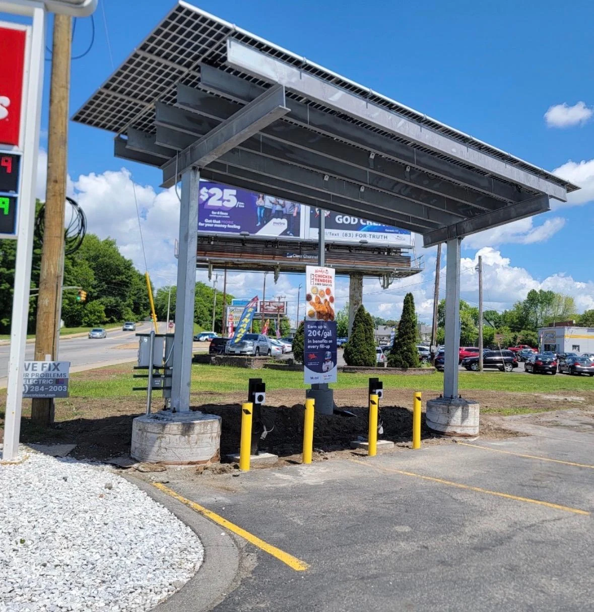 A galvanized steel solar canopy with glass panels over two parking bays, with charging posts behind yellow bollards.