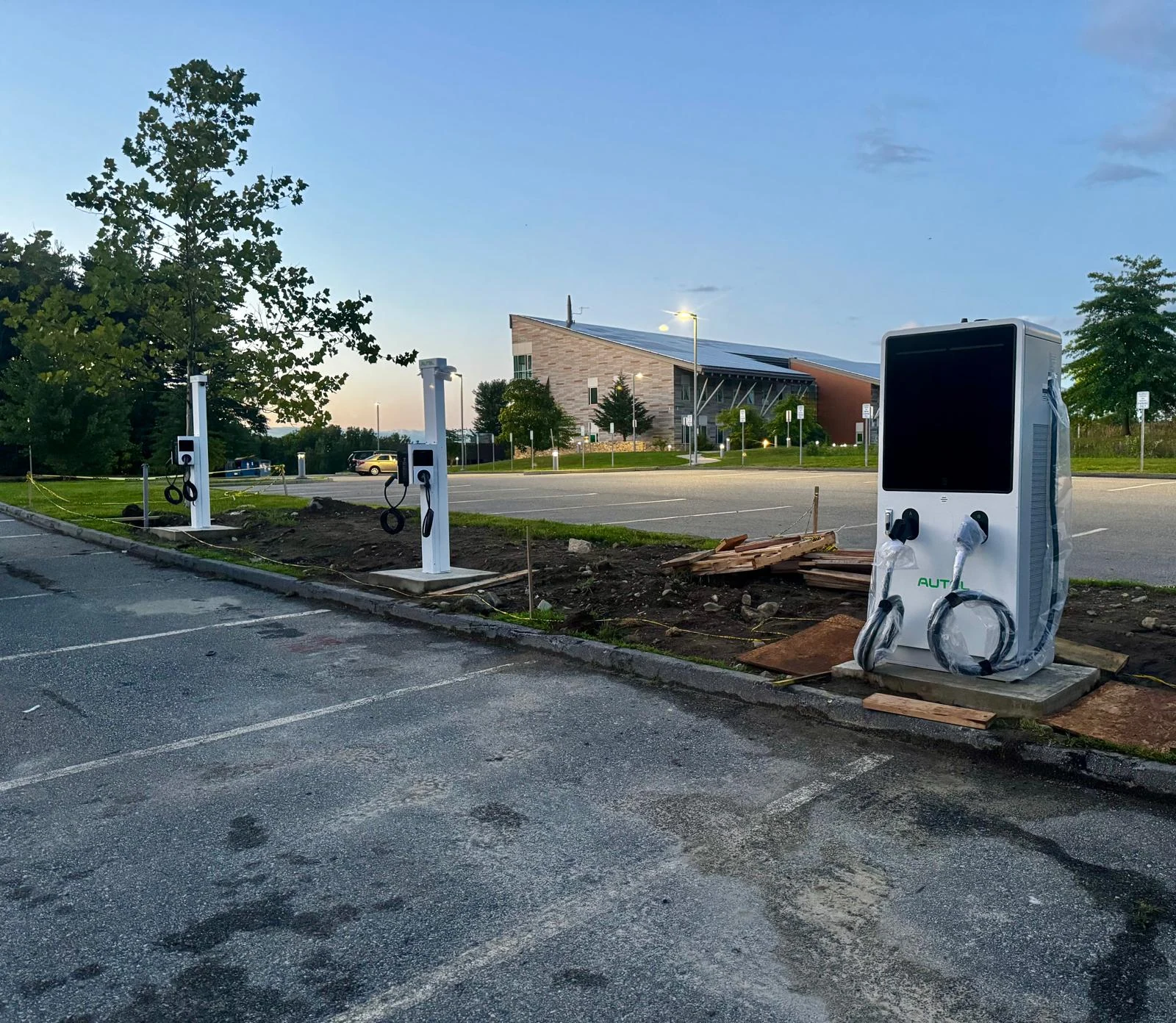Two white Level 2 charging pedestals and a DC fast charger at dusk in front of a building with a solar roof.